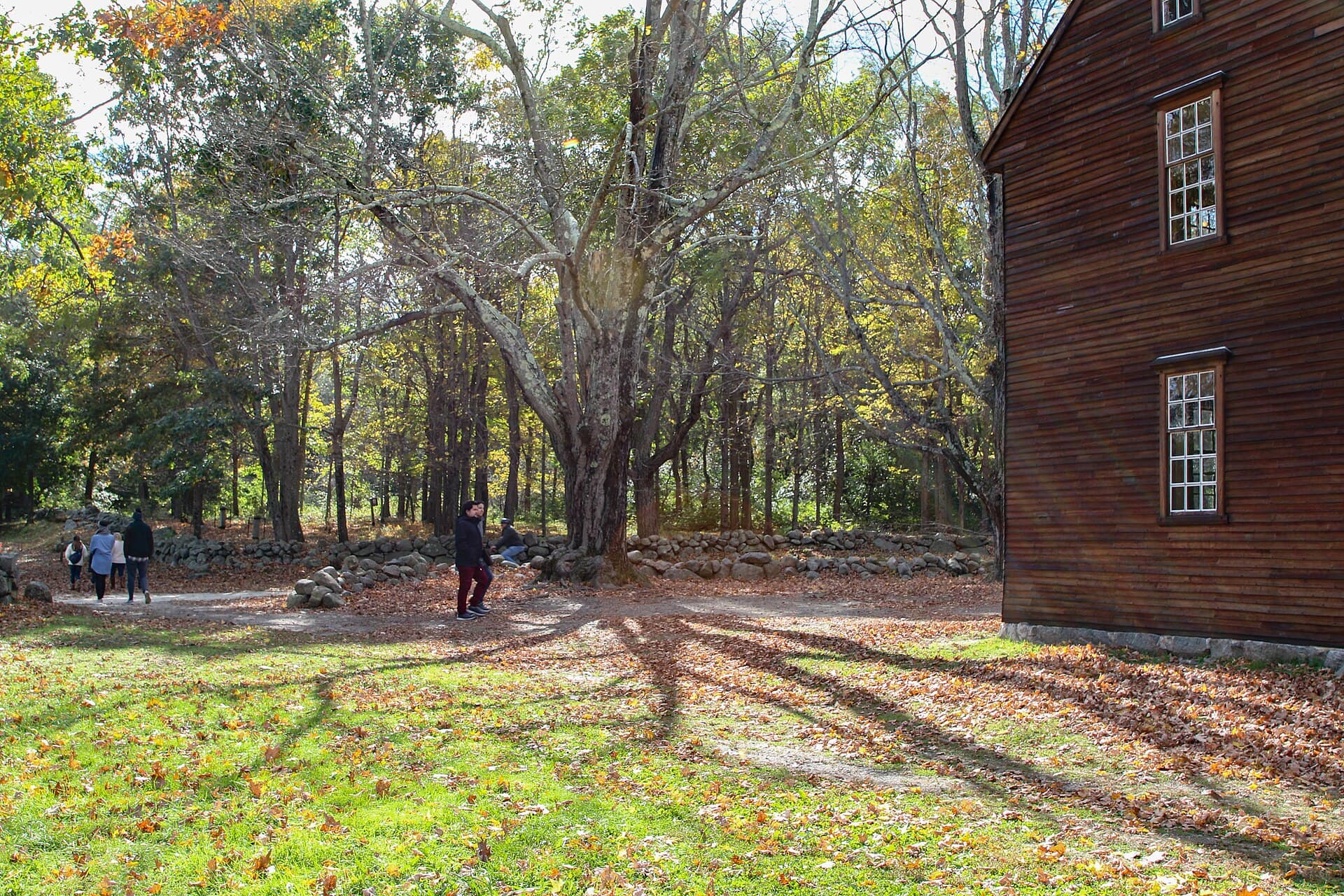 Walkers on the packed dirt Battle Road in autumn, running between fieldstone walls and bare trees, with the clapboard end of Hartwell Tavern at the right
