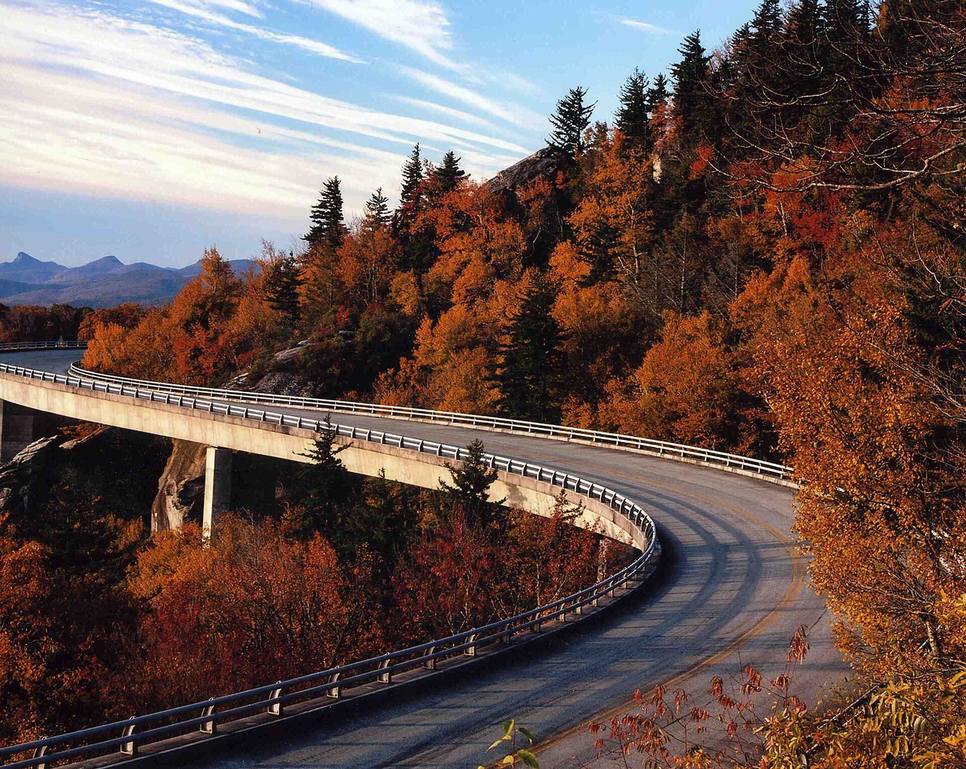 The Linn Cove Viaduct curving along a mountainside in full autumn color, its concrete deck threading between forested slopes with layered blue ridges receding into the distance under a pale sky