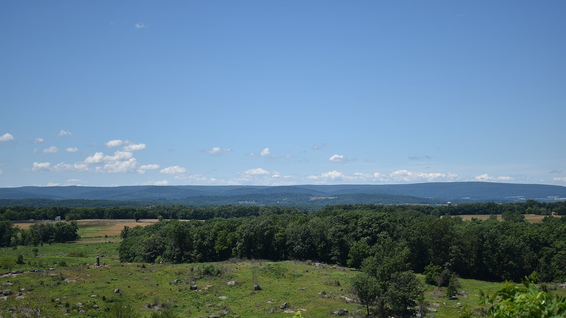 A wide view west from Little Round Top over rolling battlefield farmland and woodland toward distant blue ridges, under a pale sky with scattered clouds