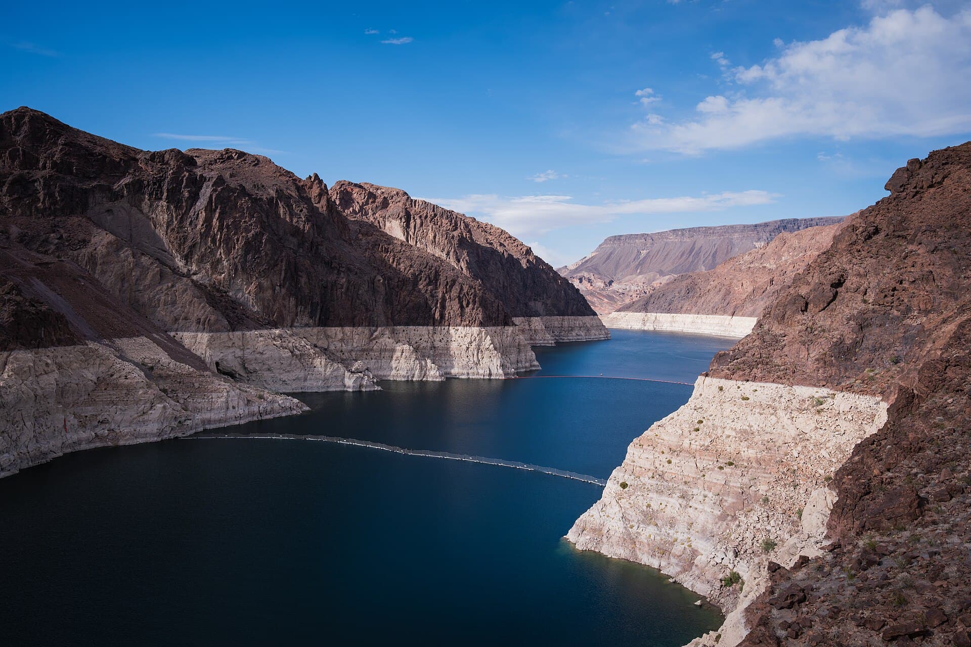 Lake Mead in Black Canyon under a deep blue sky, the dark water flanked by a brilliant white mineral band forty vertical feet high on both canyon walls where the reservoir used to stand, with a floating safety boom strung across the channel