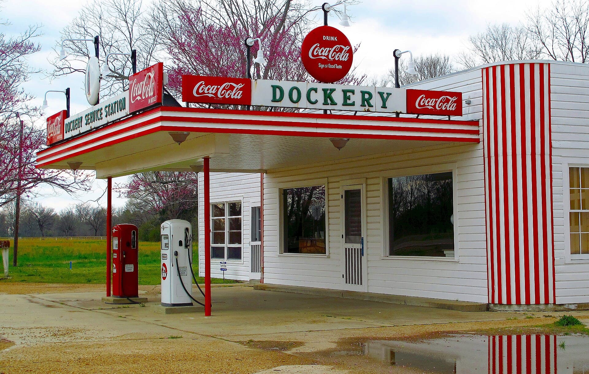 The restored Dockery Farms service station, a white clapboard building trimmed in red with Coca-Cola signs and a green DOCKERY sign along its canopy, two vintage fuel pumps standing under the awning and a redbud tree in flower behind it