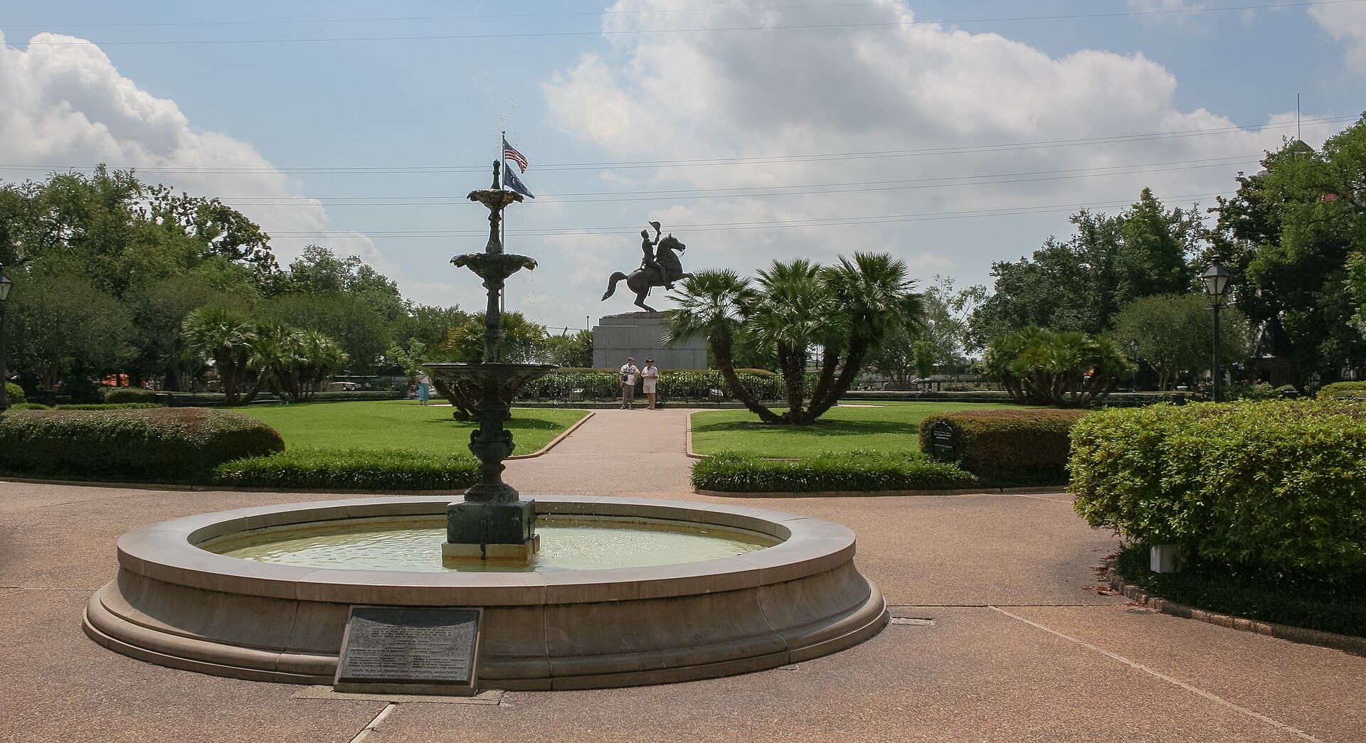 Jackson Square in bright sun, a tiered cast iron fountain in the foreground and the bronze equestrian statue of Andrew Jackson rearing on a granite pedestal beyond it, framed by clipped hedges, lawn and palms under high cloud