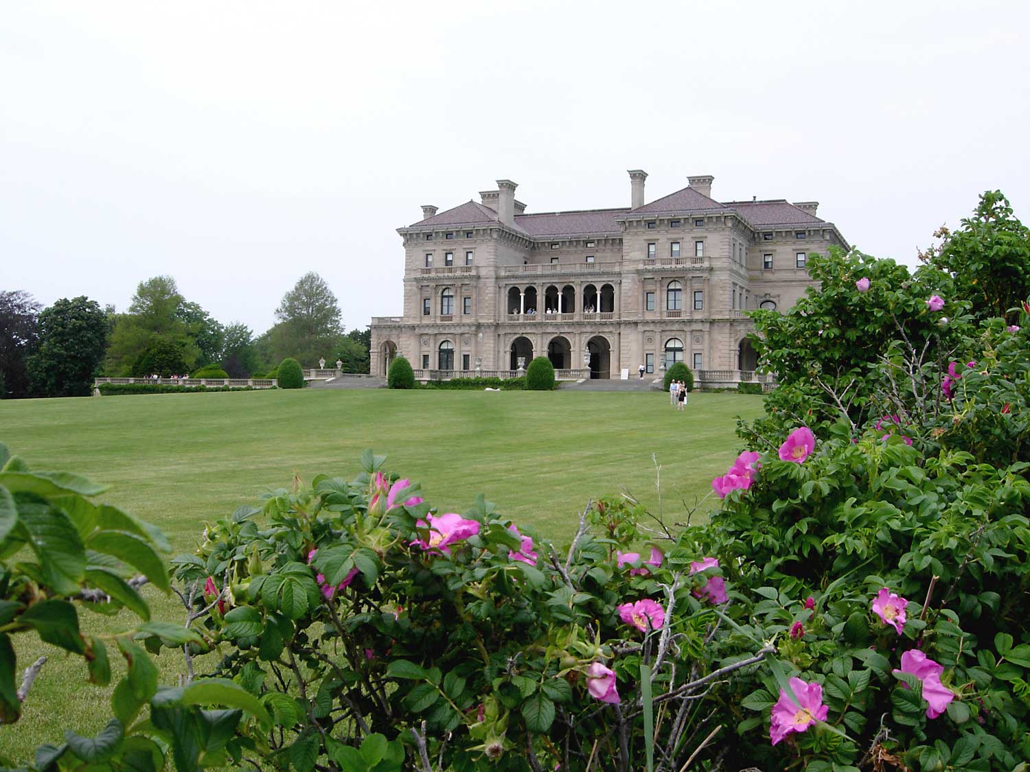 The ocean front of The Breakers seen across its wide lawn in summer, tiers of arched limestone loggias under a pale sky, with pink beach roses flowering in the hedge in the foreground