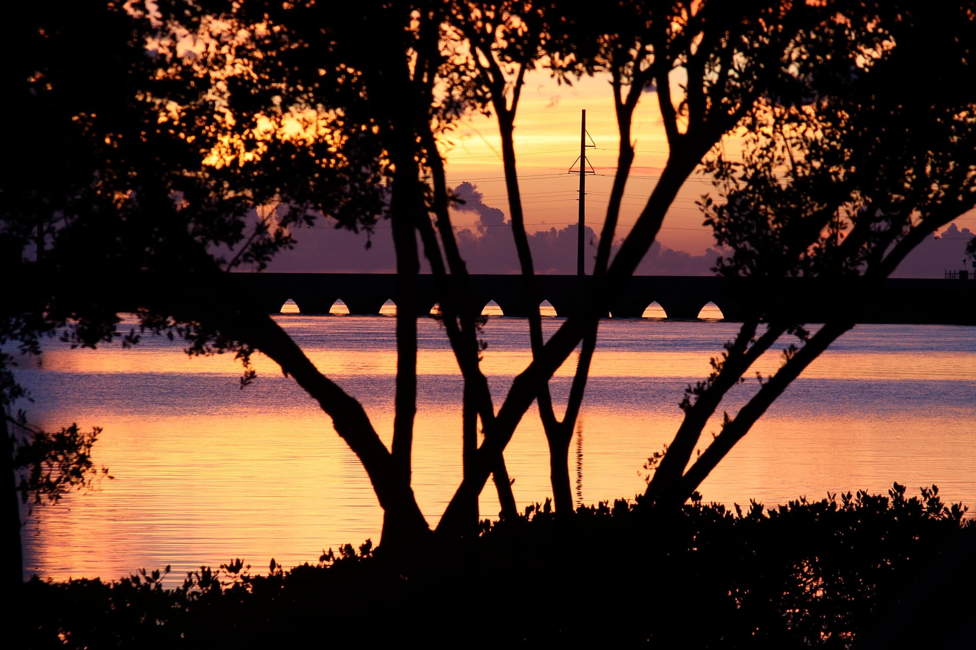 The Overseas Highway bridge near Duck Key at sunset, a low concrete span stretching toward the horizon over calm water on both sides, silhouetted power poles running alongside it under a pastel orange and pink sky