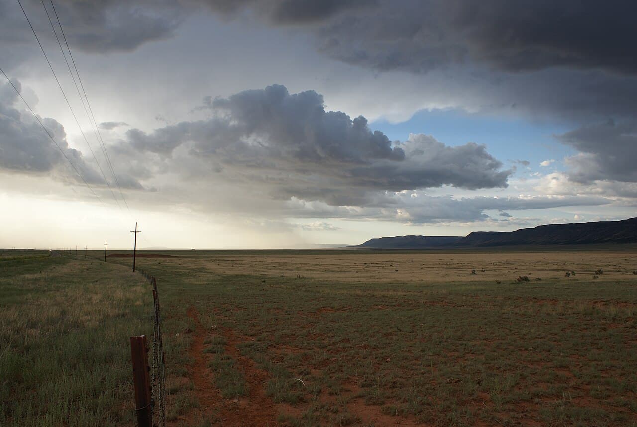 A dark storm cloud over open high desert grassland along old Route 66 near Seligman, power lines following the road toward distant flat-topped mesas under a partly lit sky