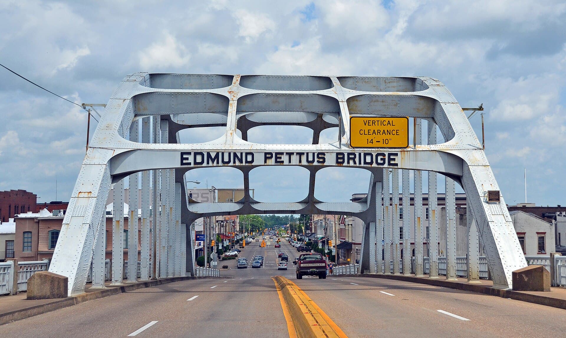 The steel arch of the Edmund Pettus Bridge seen head on from the roadway, the bridge's name in raised letters across the span, with Selma's Broad Street running away beyond it