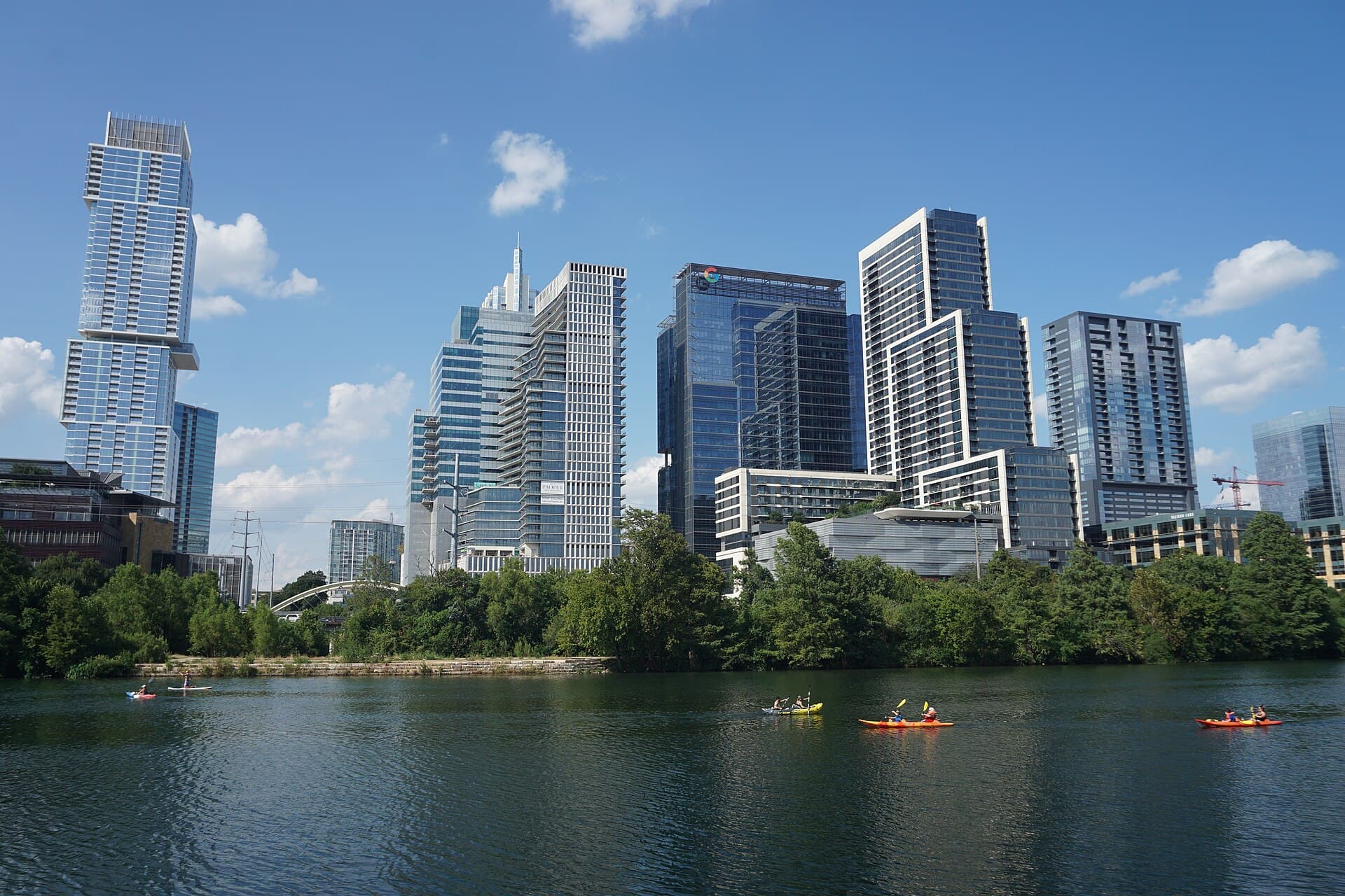 Downtown Austin's glass and concrete towers rising behind a wooded bank, seen across Lady Bird Lake on a bright summer day with kayakers and a paddleboarder on the green water in the foreground