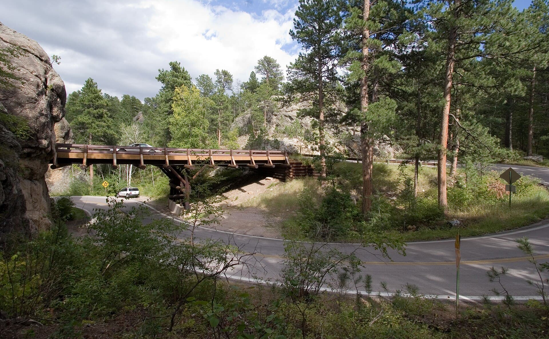 A timber pigtail bridge on Iron Mountain Road carrying a car over the same highway it just left, the road looping back beneath it through ponderosa pine and grey granite