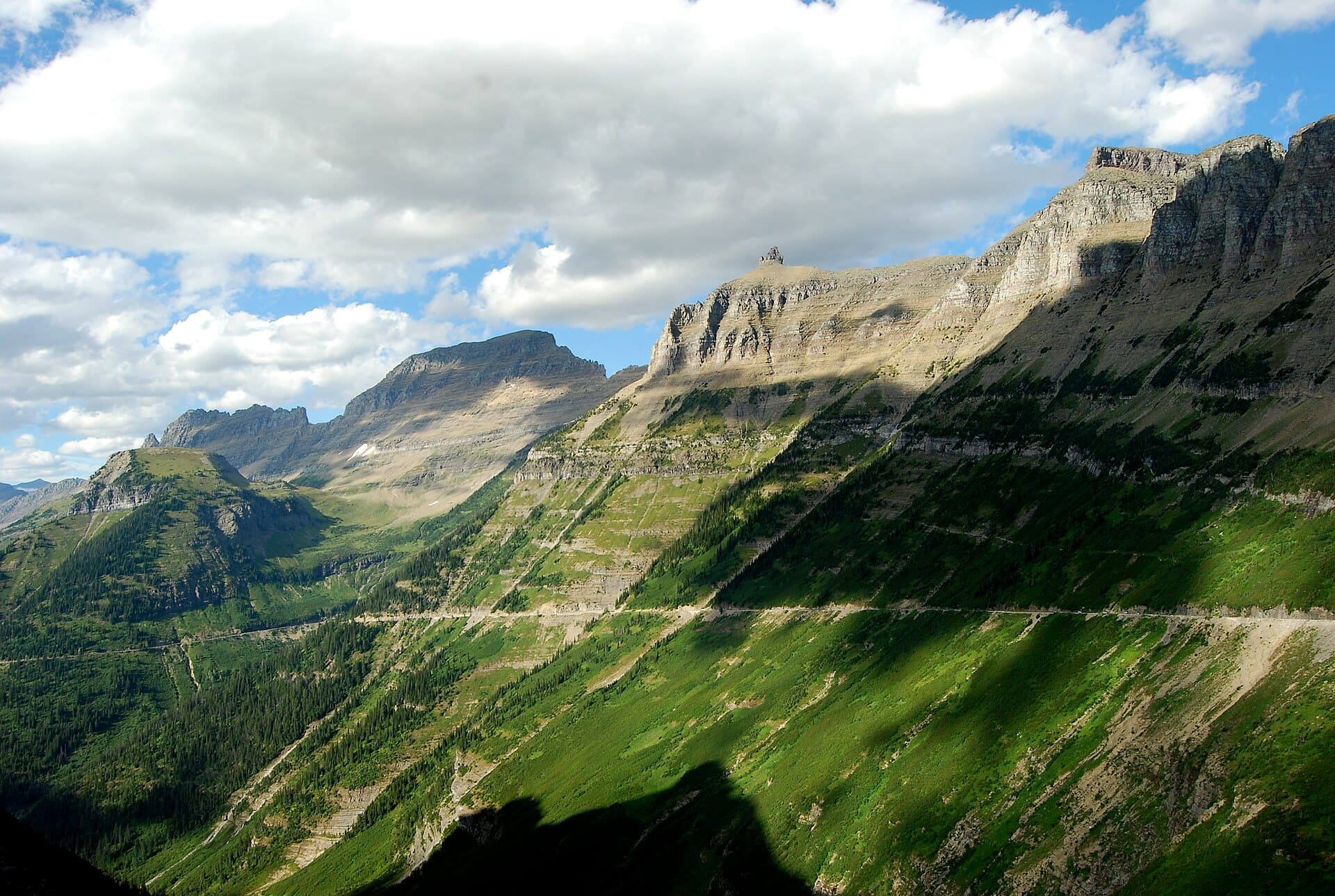 The Going-to-the-Sun Road seen from across a valley as a thin level line cut into a long green mountainside, rising toward tan cliffs and rock summits under a blue sky full of white clouds