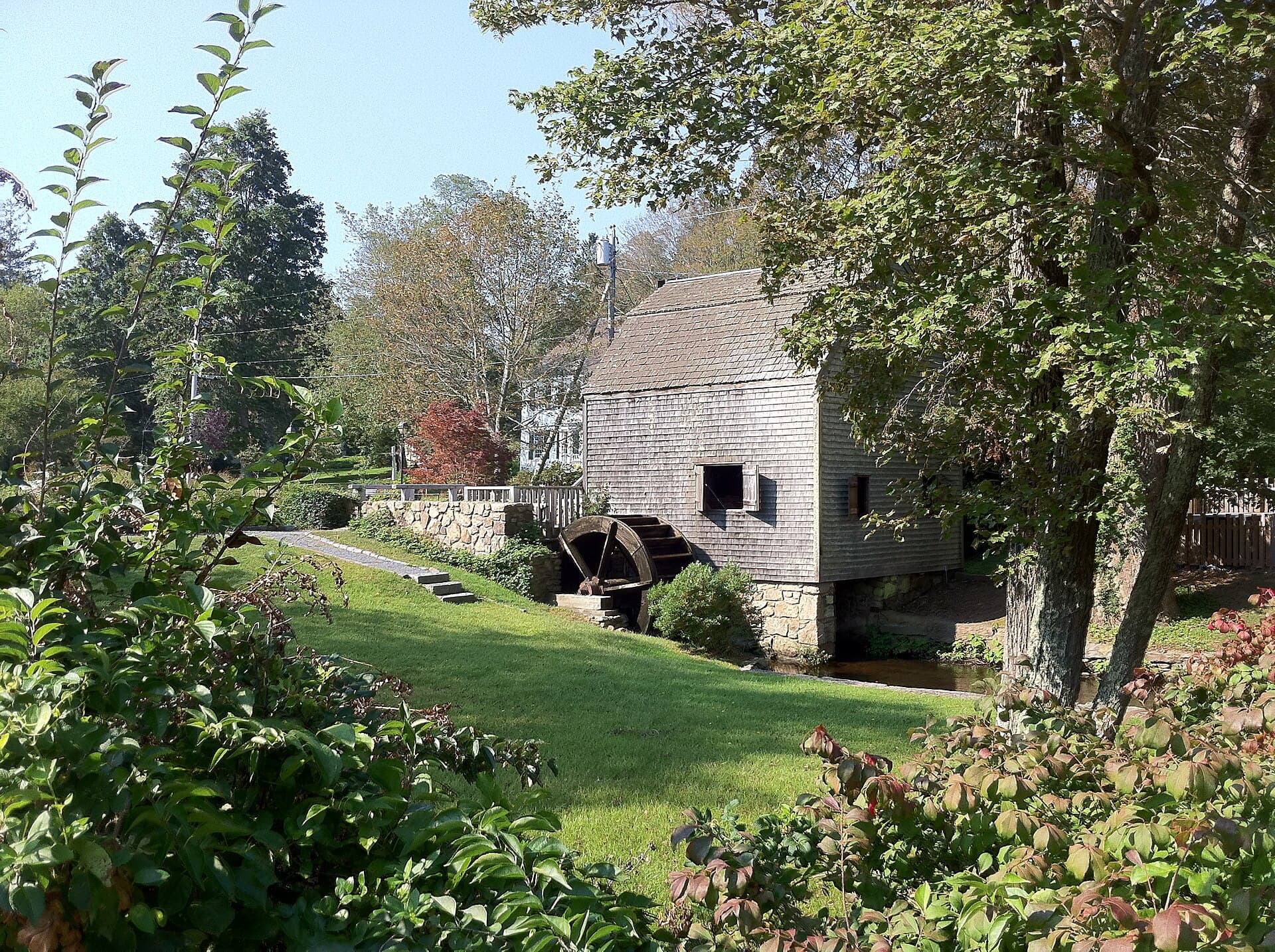 The shingled Dexter Grist Mill in Sandwich beside its wooden waterwheel and millrace, with a lawn running down to the water and trees crowding in on both sides