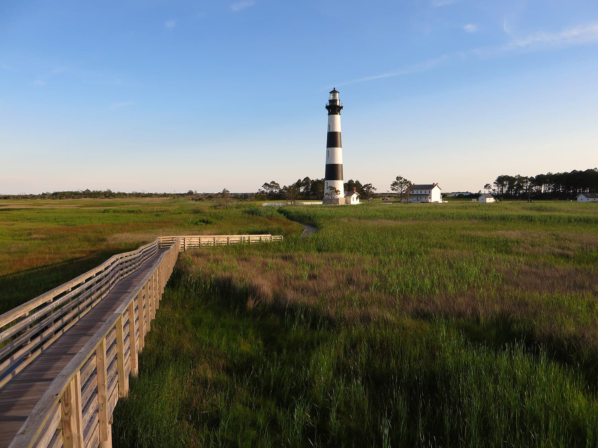 Bodie Island Lighthouse in black and white bands, seen across salt marsh from a wooden boardwalk in evening light