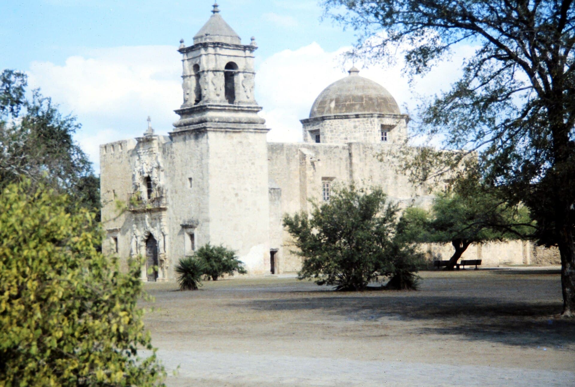 The limestone church at Mission San José seen across the open compound, its carved portal and single bell tower to the left of a low dome, framed by mesquite