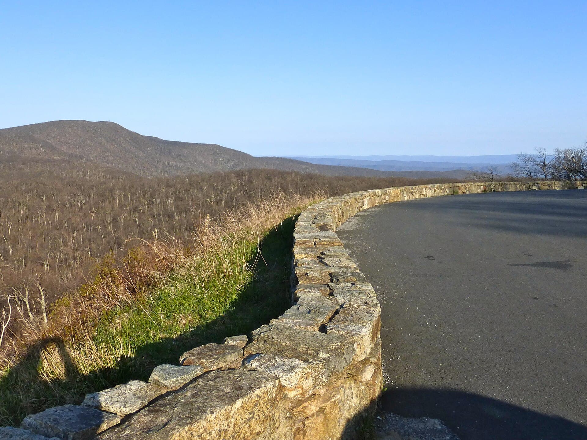 A dry-laid stone guard wall curving along the edge of Skyline Drive, the empty road on the right and a steep slope of leafless early-spring forest falling away to the left, with a rounded mountain and a hazy blue horizon under a clear sky